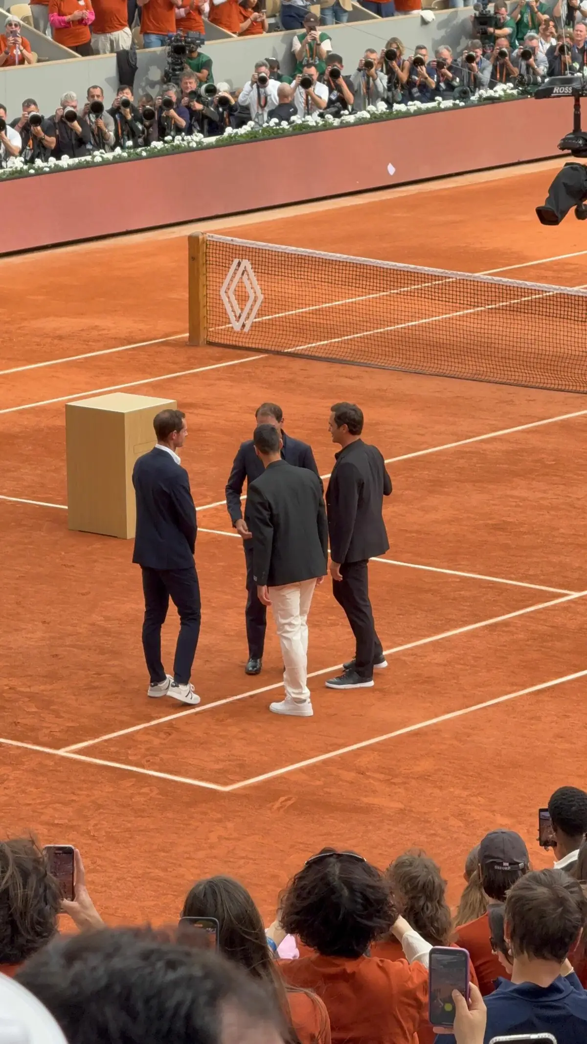 Vier Spieler in Anzügen auf dem Sandplatz von Roland Garros, dahinter die Fotografentribüne